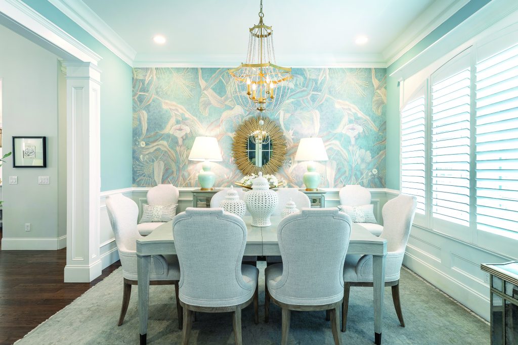 Elevate your home for the holidays with an elegant dining room featuring light-colored chairs, a decorative mirror, and floral wallpaper. A chandelier sparkles above the table, while windows with shutters flood the space with natural light.