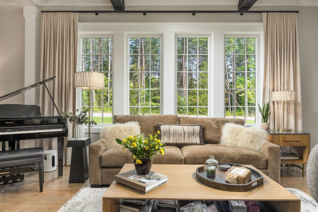 A cozy living room with a brown sofa, fluffy pillows, and a coffee table with flowers and decor. A piano is on the left, tall windows with curtains overlook a green yard.